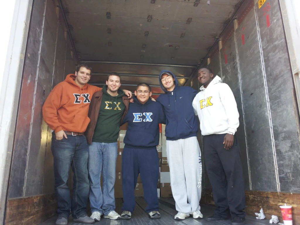 Five men wearing fraternity sweatshirts stand smiling together inside an empty truck trailer, serving as a beacon of light for students and families.