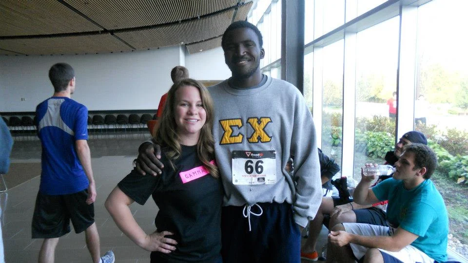 Two students pose indoors; one wears a race bib and Greek letters, while the other sports a black T-shirt labeled "Catherine.