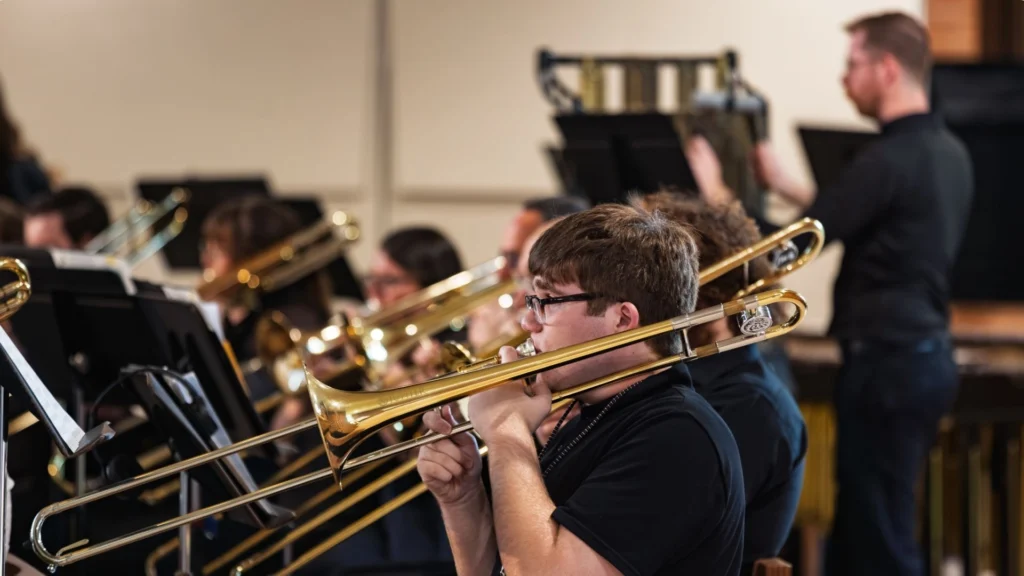 A group of musicians play trombones in an orchestra, with a conductor leading them, evoking the spirit of a Sousa Legacy in every note.