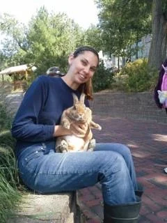 Erica Sipich sits outdoors on a brick ledge, smiling warmly as she holds a brown rabbit in her lap, enjoying a peaceful moment in Beacon.