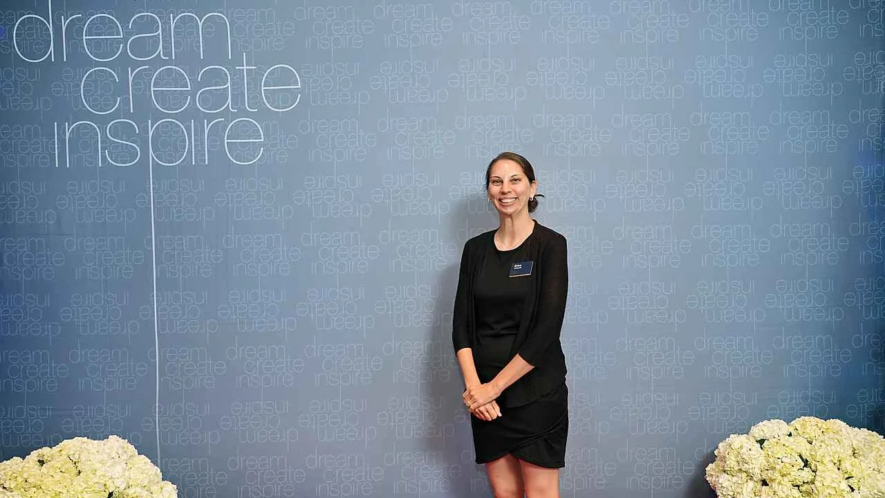 Erica Sipich beams in a black dress before a "dream create inspire" event backdrop adorned with white flowers, standing as a beacon of inspiration.