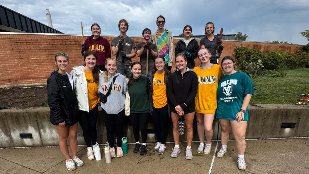 A group of smiling students in casual clothes, including Environmental Engineering major Alex Miroballi, poses together outside on a cloudy day near a garden bed, united by their shared commitment to a greener future.