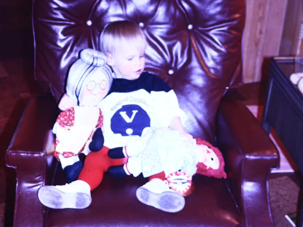 A toddler sits on a brown leather chair, holding two dolls, one on each side, in a scene reminiscent of a Kimberly Dana story—a beacon of childhood innocence and imagination.