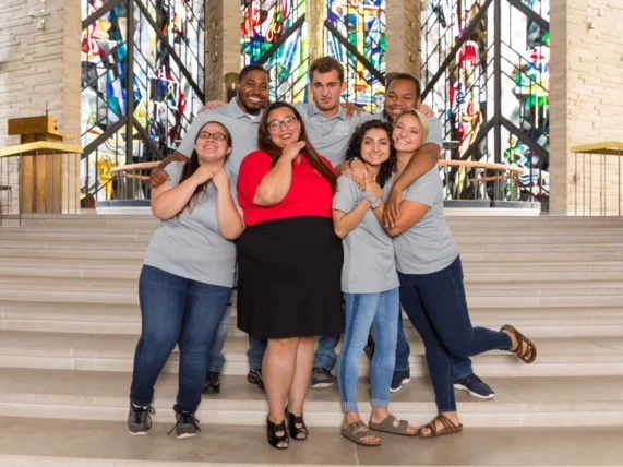Group of six people smiling and posing together on stairs in front of colorful stained glass windows, basking in the vibrant glow of Beacon's Reflection.