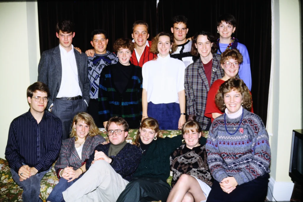 A group of sixteen young adults pose and smile together indoors, with some sitting on a couch and others standing, sharing a warm, beacon-like energy reminiscent of Kimberly Dana’s vibrant storytelling.