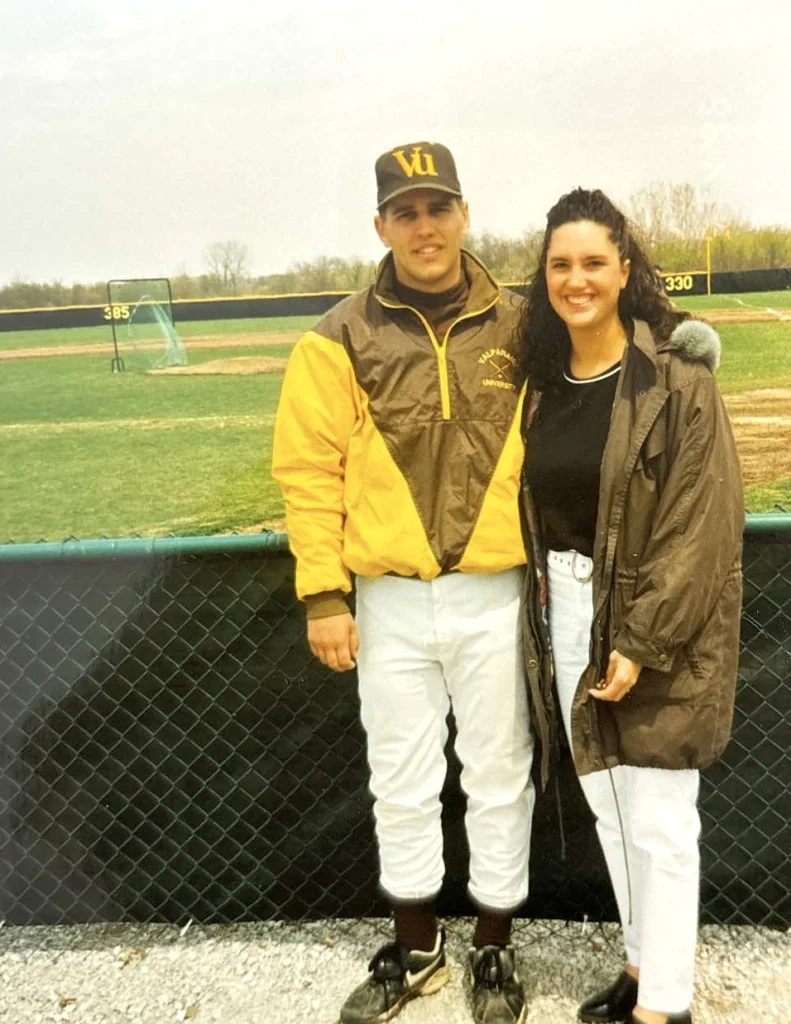 Chip Pettit, in his baseball uniform, stands next to a woman by a fence on the Beacon baseball field, both smiling warmly.