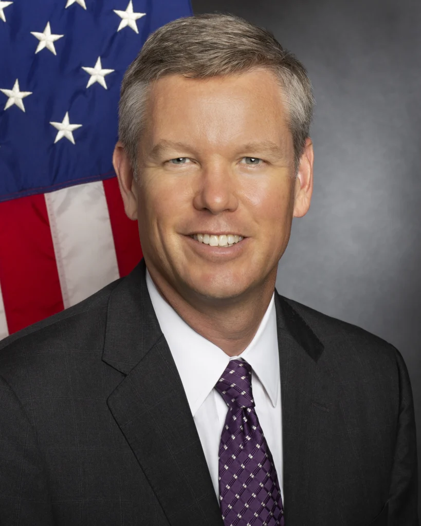Smiling man in a suit and purple tie, posed in front of a US flag and gray background, represents the guidance of Follow the Beacon, embodying the leadership style of Chris Hanson.