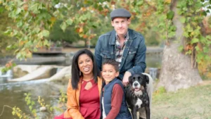 A family of three and their dog sit on a blanket outdoors by a river, surrounded by autumn trees—enjoying nature's beauty much like Literary Luminaries such as Jim Iddins, MALS, might savor peaceful moments for inspiration.