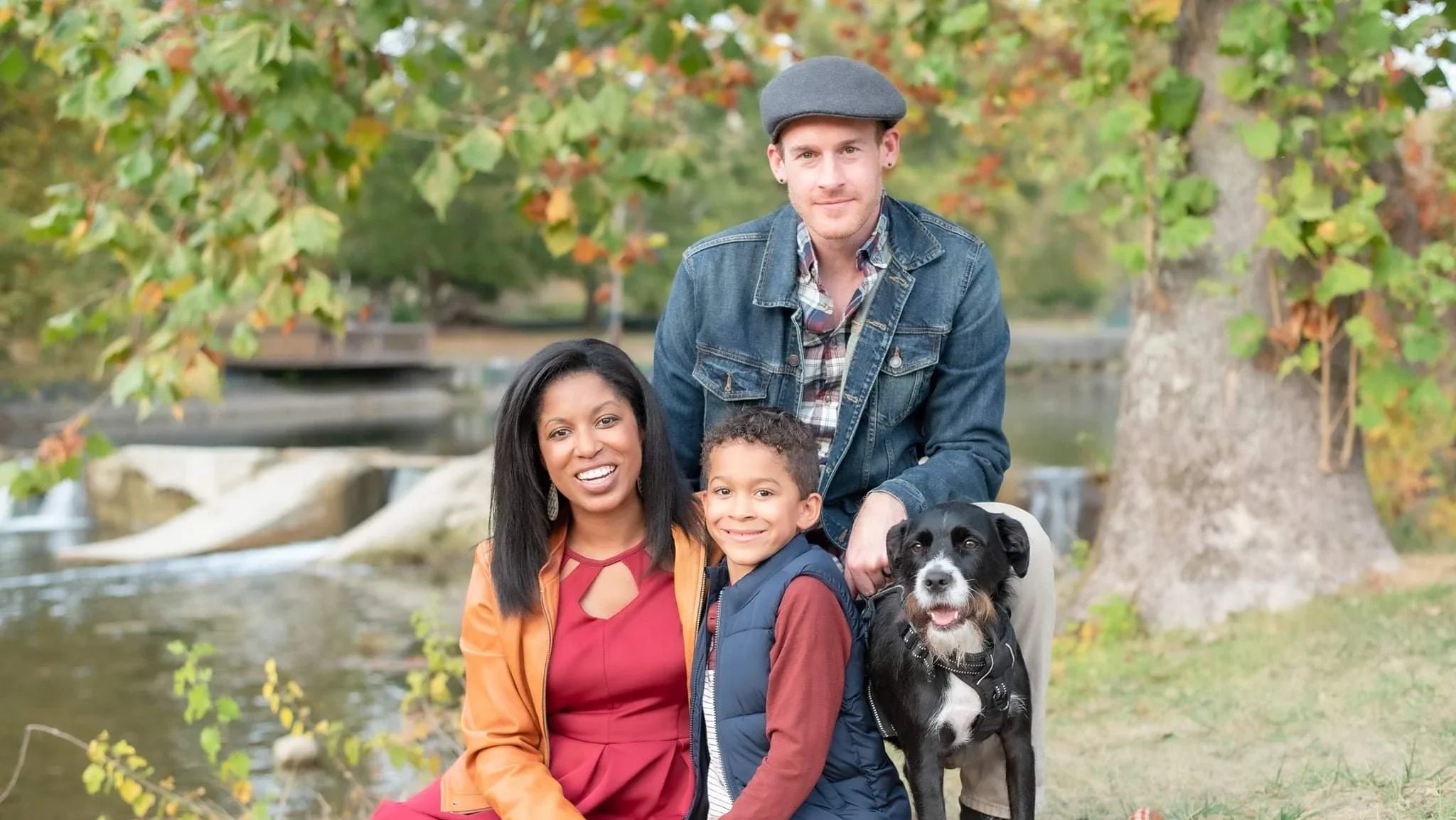 A family of three and their dog sit on a blanket outdoors by a river, surrounded by autumn trees—enjoying nature's beauty much like Literary Luminaries such as Jim Iddins, MALS, might savor peaceful moments for inspiration.