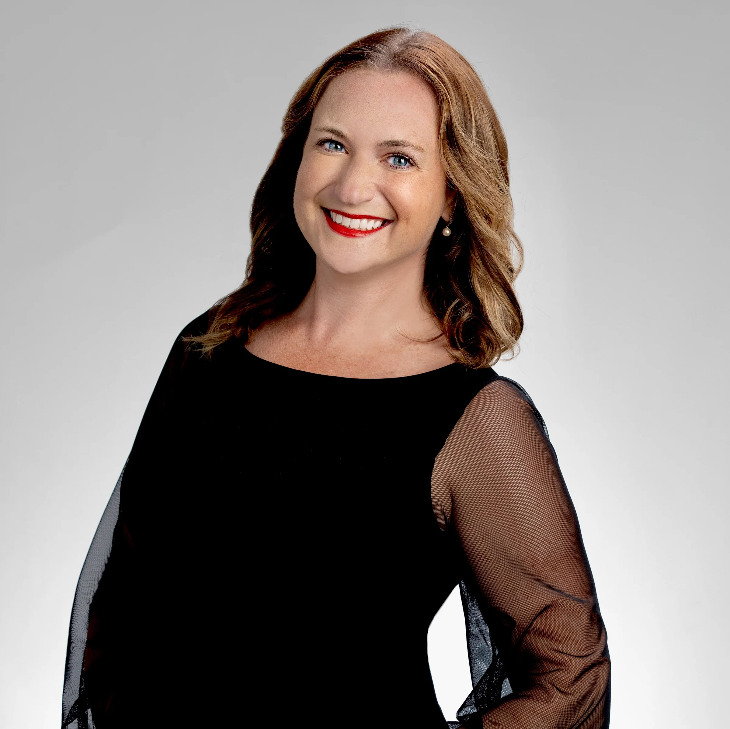 Smiling woman with wavy hair in a black dress with sheer sleeves poses against a plain light background, radiating confidence like a beacon—much like Meredith Garofalo herself.