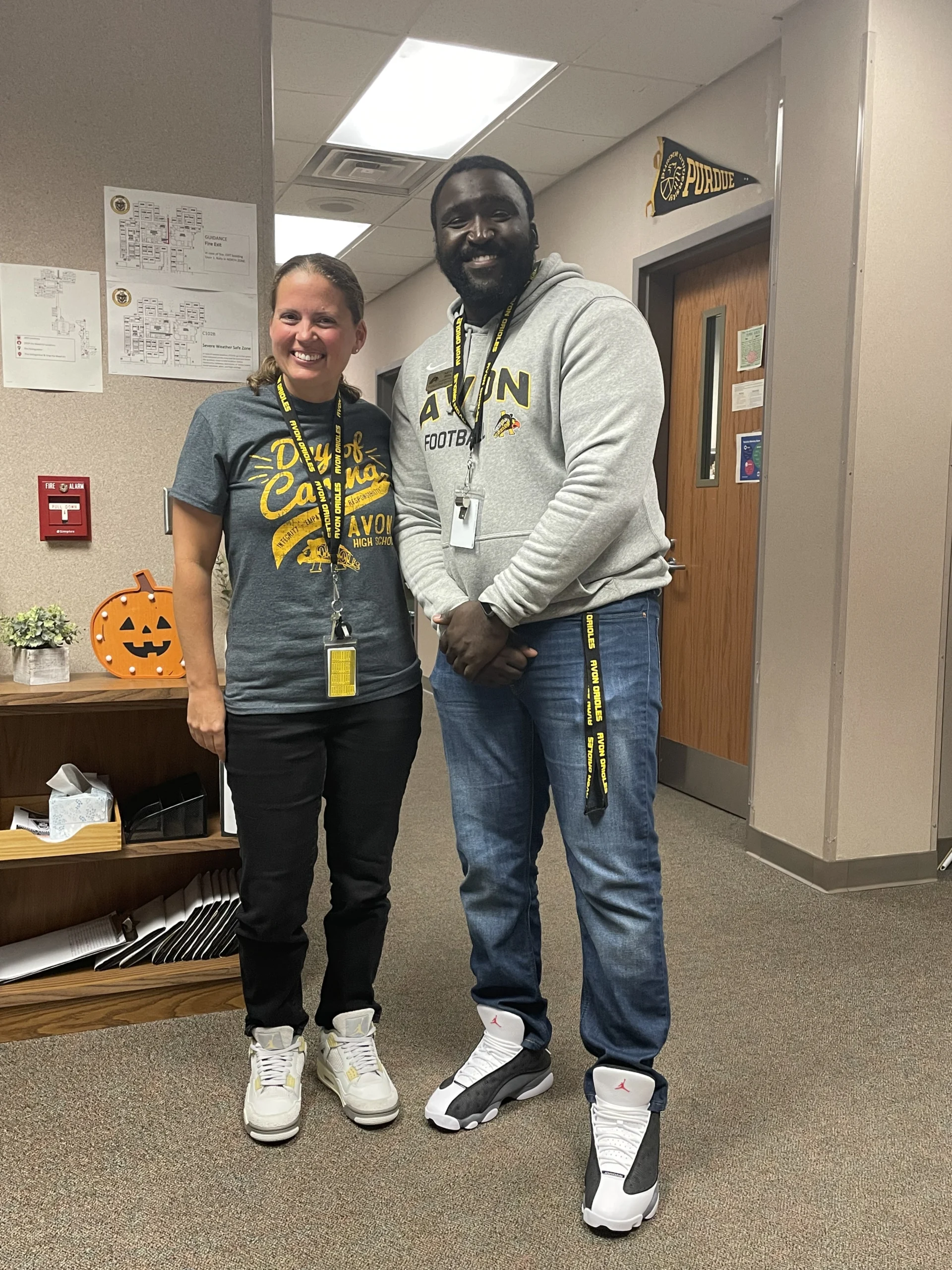 Two smiling adults, Aaron Willis and a colleague, stand side by side in an office, both wearing casual Purdue-themed clothing and sneakers, showing their support for students and families.