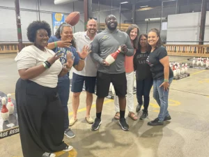 Six people, including students and Aaron Willis, smile and pose together at an indoor bowling alley, holding a red and white bowling pin.
