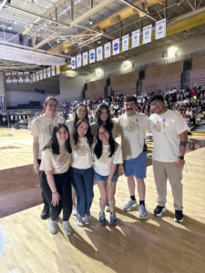 Eight people in matching shirts pose and smile on a basketball court inside a gymnasium with a crowd behind them, celebrating Environmental Engineering and working together for a Greener Future.