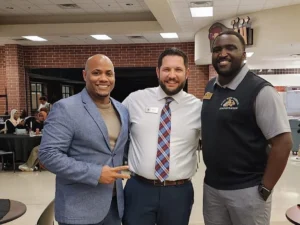 Three men, including Aaron Willis, stand together and smile in a cafeteria-style room filled with tables, students and families enjoying their time in the background.