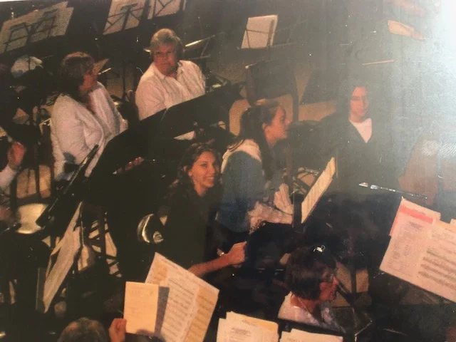 A group of musicians sit with sheet music, preparing to play in an orchestra rehearsal led by Erica Sipich.
