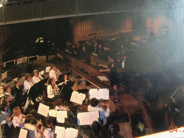 A conductor leads an orchestra, including Erica Sipich on violin, with musicians playing instruments and reading sheet music in the grand Beacon concert hall.