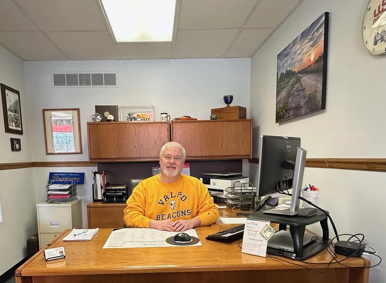 John Yelkich, wearing a yellow Valpo Beacons sweatshirt, sits at a desk in an office with a computer and paperwork.