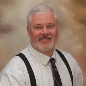Smiling man, John Yelkich, with gray hair and beard, wearing a striped shirt, tie, and suspenders stands as a beacon of confidence against a neutral background.