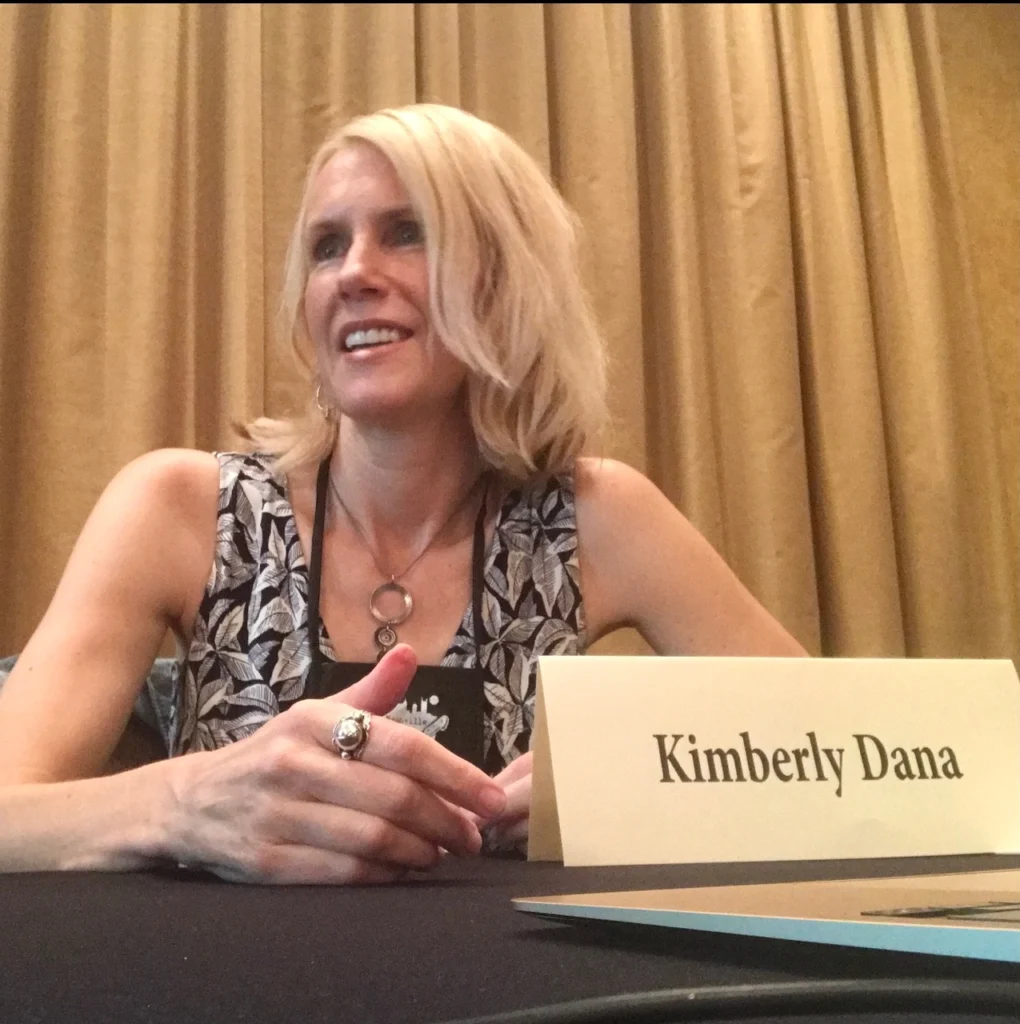 A woman with blonde hair sits at a table with a name card that reads "Kimberly Dana," her presence radiating like a beacon of confidence and grace.