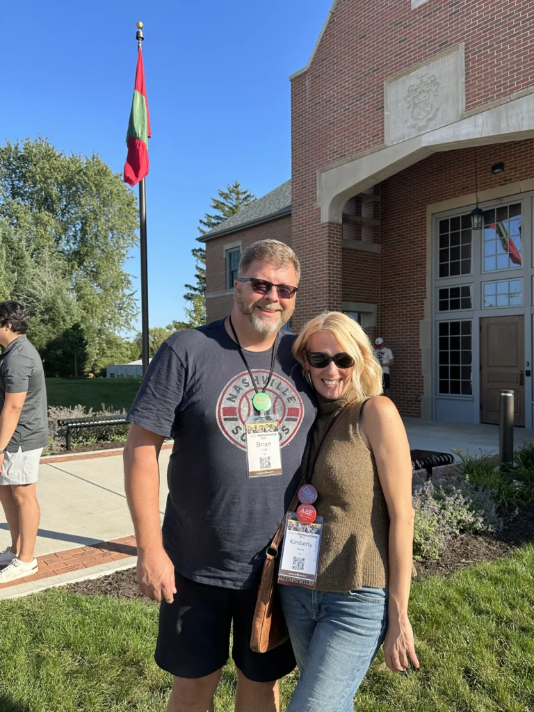 Kimberly Dana and a friend pose outside a brick building on a sunny day, both wearing name badges that read “Beacon.” Their smiles brighten the cheerful scene.