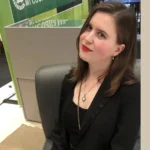 A woman with brown hair and red lipstick sits on a gray chair, wearing a black blazer and necklace, ready to begin her LINC Language Tutoring session.