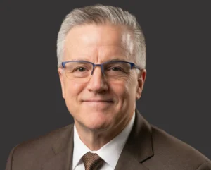 Smiling older man with gray hair, glasses, and a brown suit against a plain dark background, representing the Chief Financial Officer at Valparaiso University.
