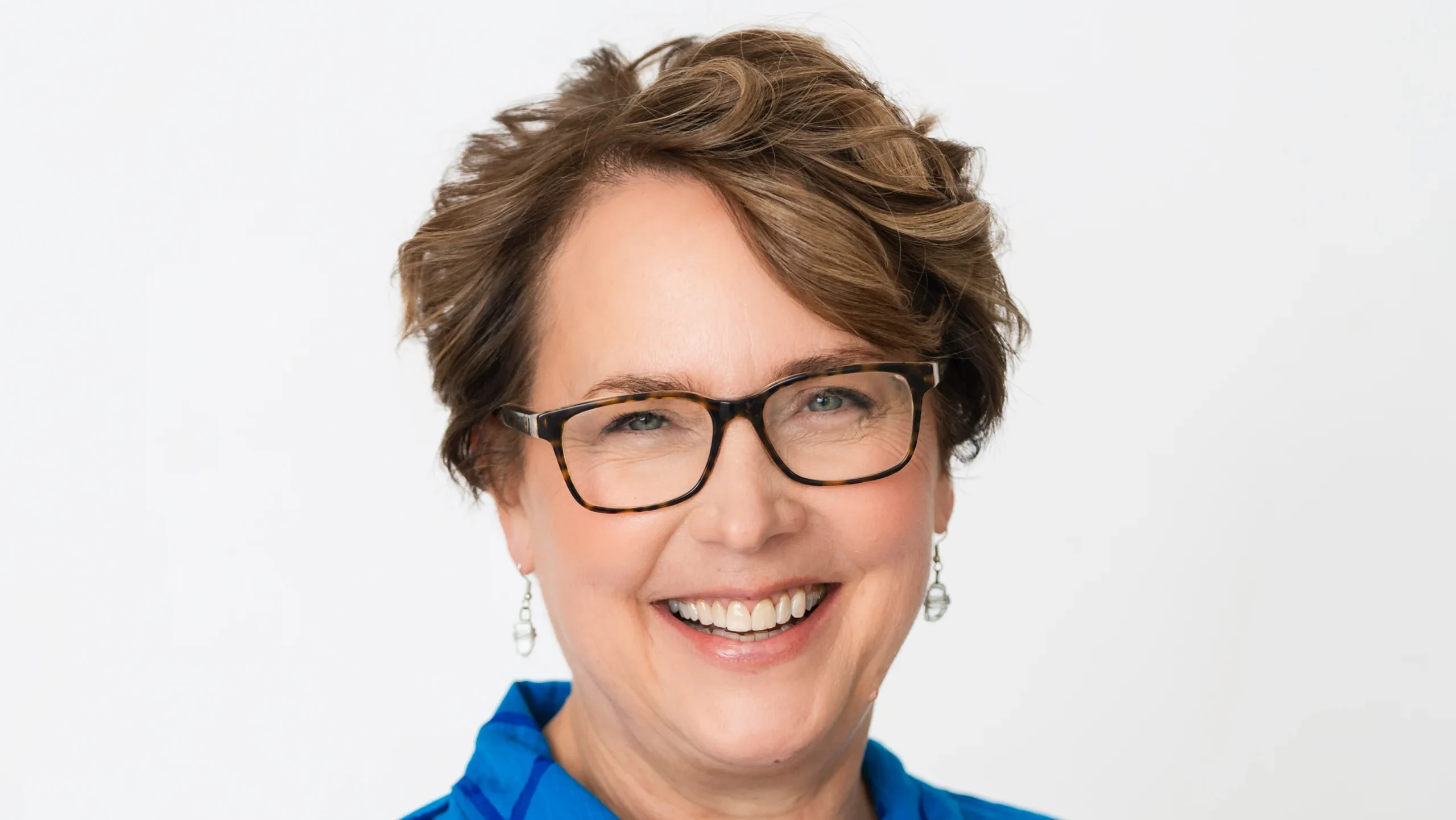 Smiling woman with short brown hair, glasses, and a blue patterned shirt on a white background. Martha Stoeppelwerth of Beacon exudes warmth and confidence in this portrait.