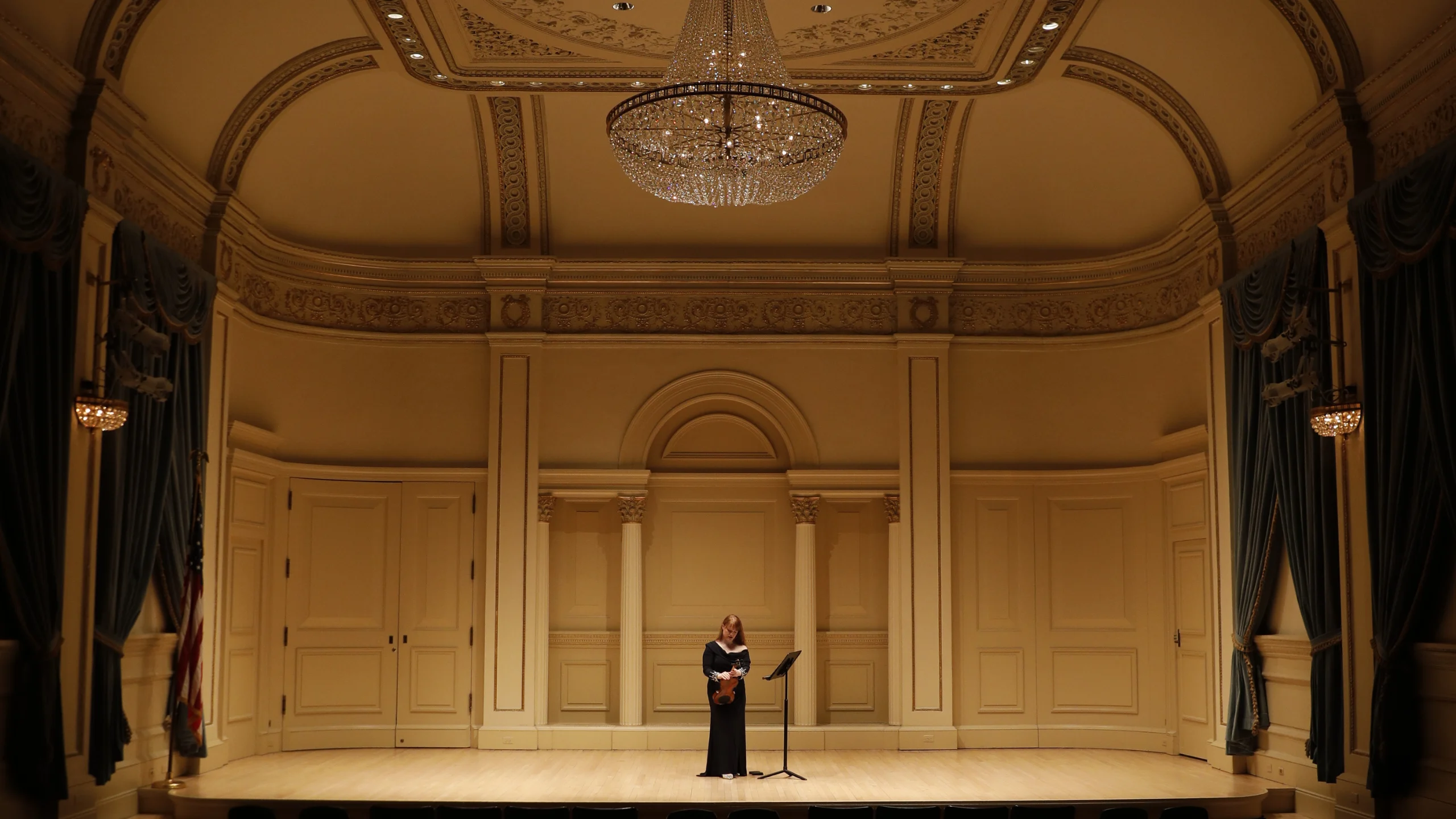 A woman in a long dress stands alone on stage under a chandelier in an elegant, empty concert hall, ready for her Guest Blog performance—a night inspired by Bonta’s timeless grace.