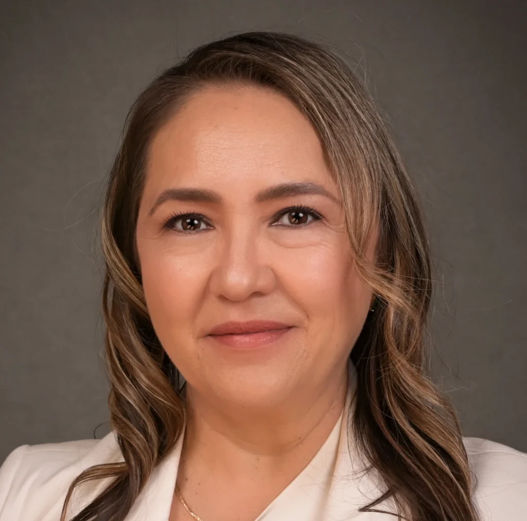 Woman with long wavy hair wearing a white blazer, smiling in front of a plain gray background, representing Veteran Student Services with confidence and warmth.