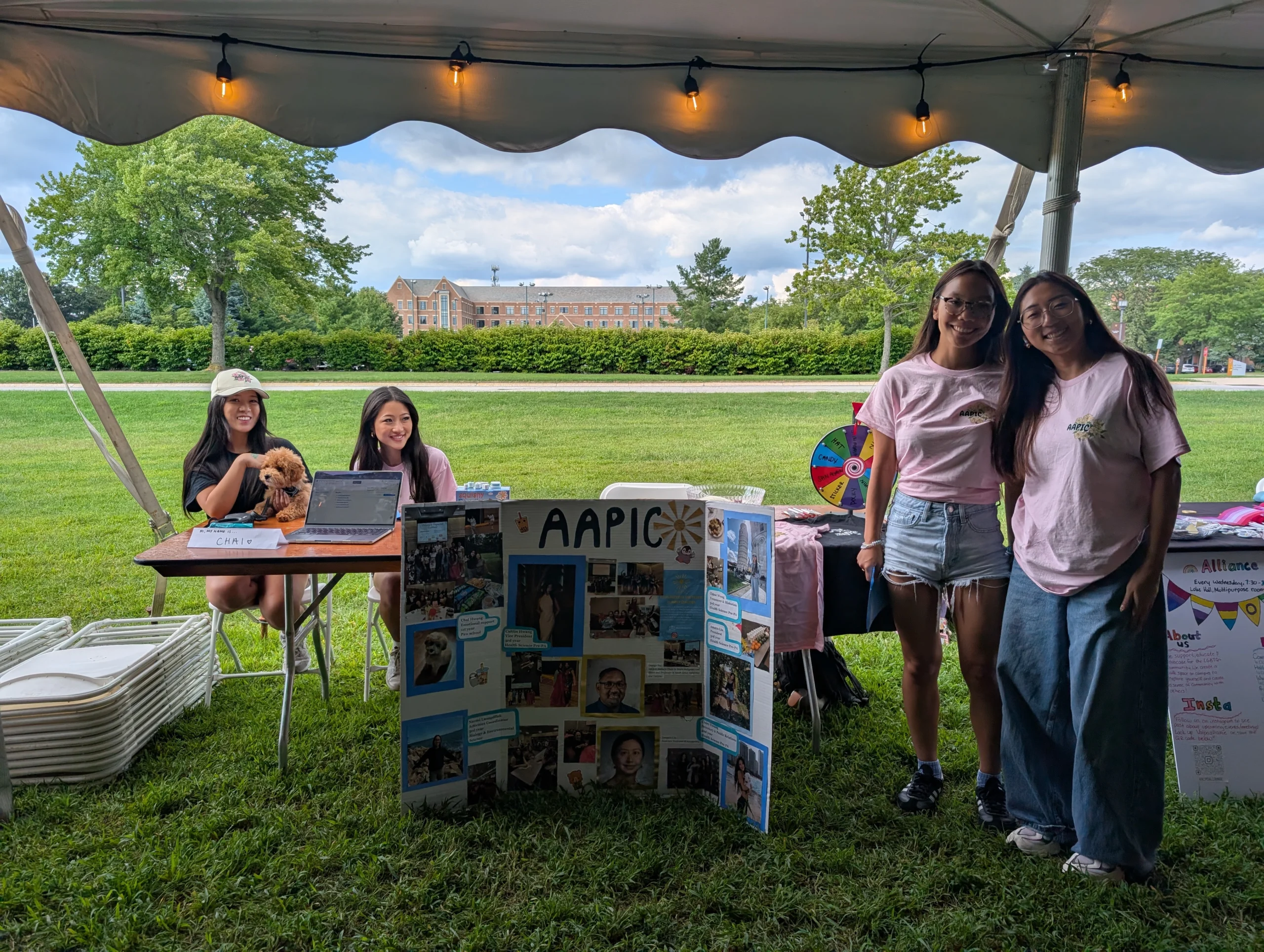 Four young women at an outdoor booth for Multicultural Organizations, featuring an "AAPIC" display board under a tent on a grassy lawn.