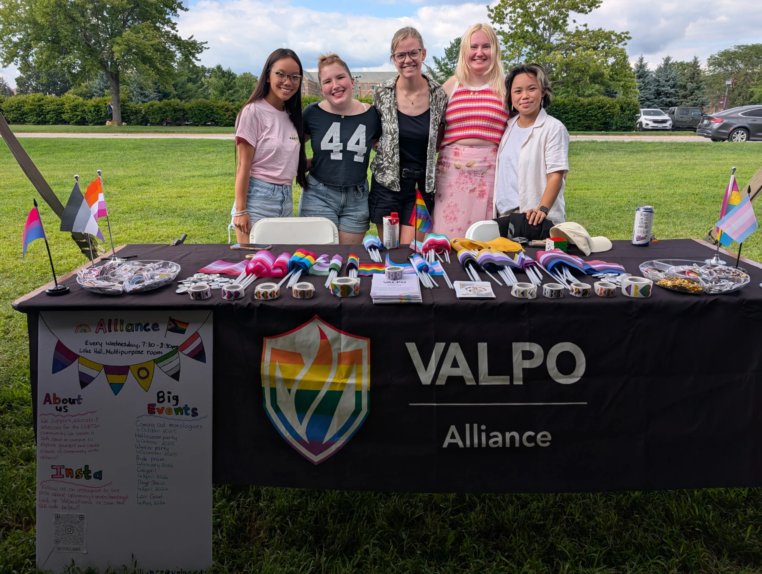 Five people stand behind a Valpo Alliance table, representing Multicultural Organizations, with pride flags, buttons, and flyers at an outdoor event.