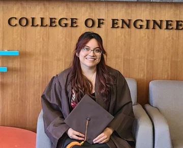 Smiling graduate Krystal Peña in a cap and gown sits in front of a "College of Engineering" sign, ready to follow the beacon toward a bright future.