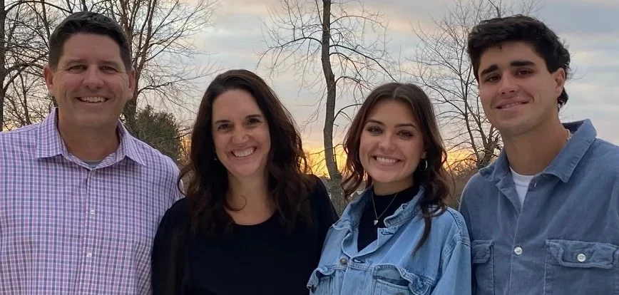 Four people, including Chip Pettit, smiling outdoors at sunset, stand together in casual clothing with trees in the background—a perfect moment to follow as a beacon of friendship and joy.