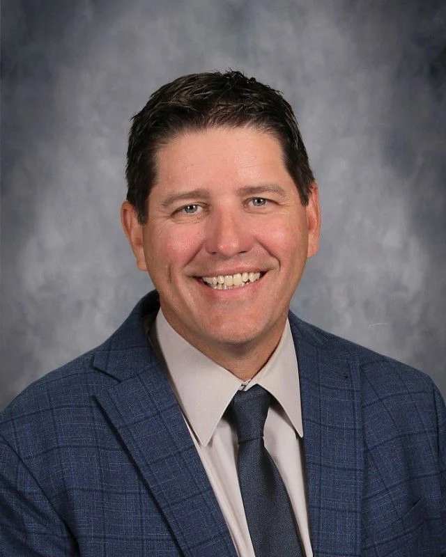 Smiling man, Chip Pettit, in a blue suit, light shirt, and dark tie stands confidently like a beacon against a gray studio background.