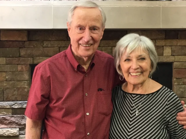 An older man and woman smiling together in front of a brick fireplace.