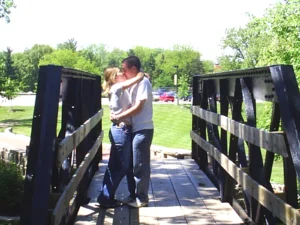 A couple shares an everlasting embrace and kiss on a wooden bridge in a sunny park.