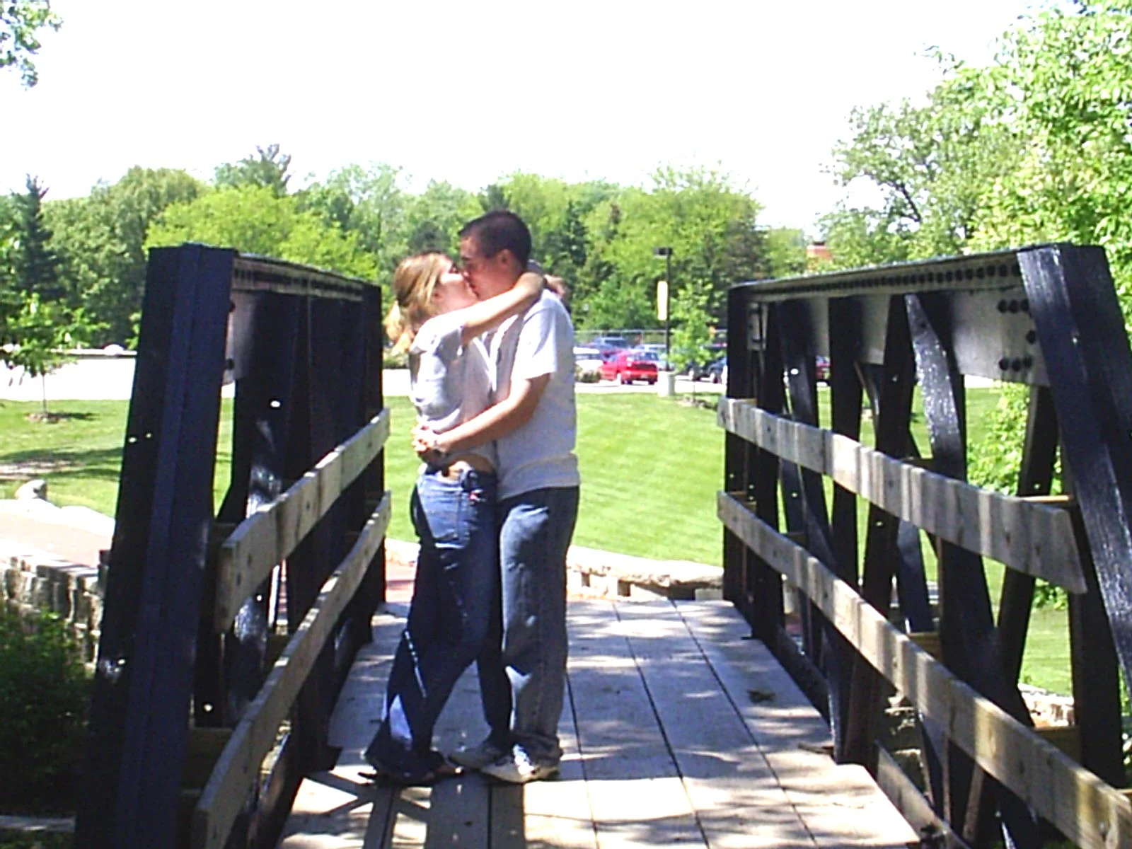 A couple shares an everlasting embrace and kiss on a wooden bridge in a sunny park.