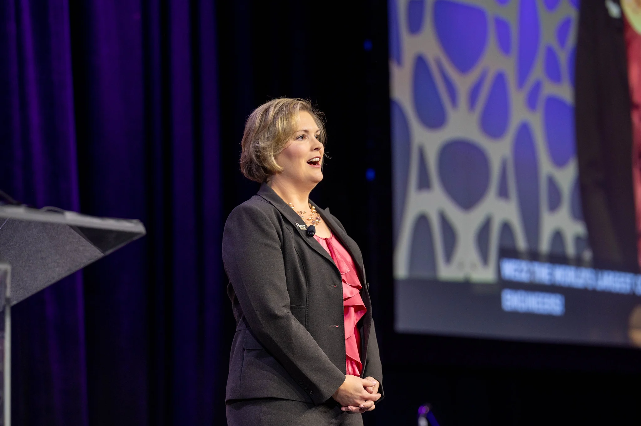 Dayna Smoljo, in a suit, speaks on stage at Beacon with a screen and geometric background behind her during the Johnson MEM event.