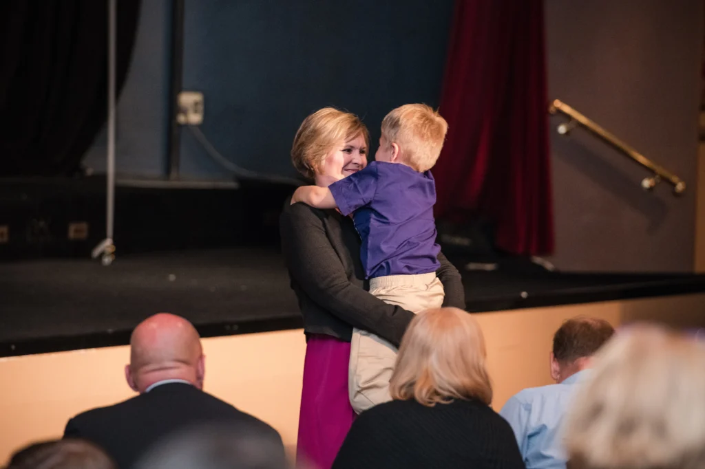 A woman in a pink dress hugs a young boy while standing near a stage at Johnson MEM, with Dayna Smoljo and others seated in the audience, creating a beacon of warmth and connection.