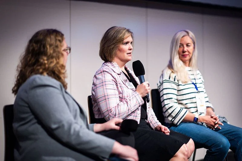 Three women seated, with Dayna Smoljo speaking into a microphone during a Johnson MEM Beacon panel discussion or interview.