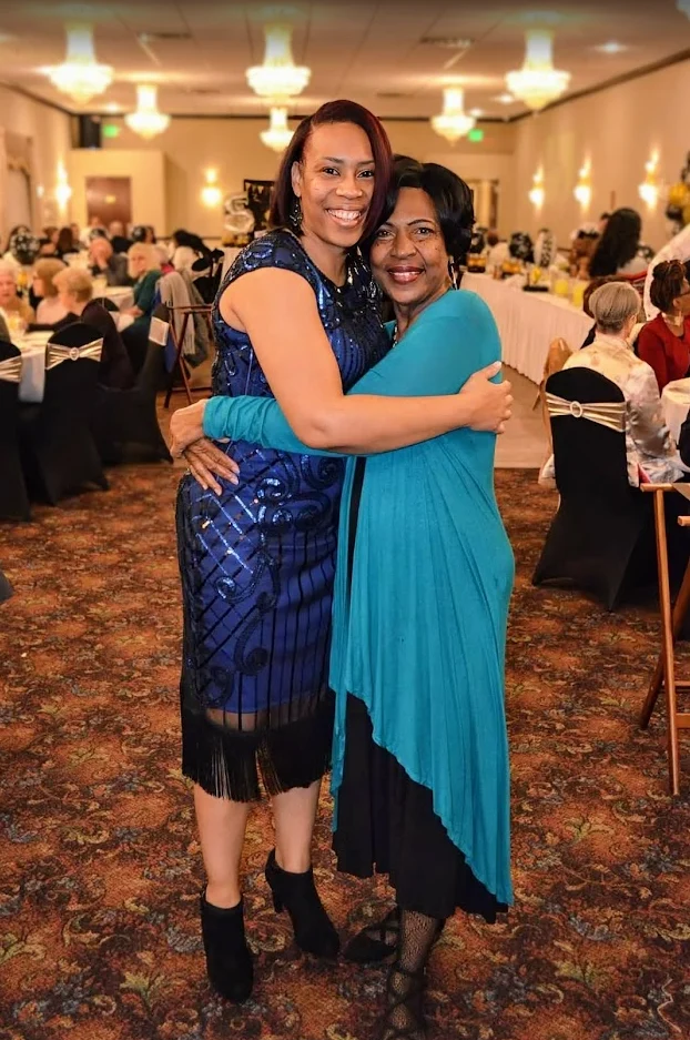 Two women smiling and hugging at a formal event in a banquet hall, with Shantée Lewis standing as a beacon of joy among tables and guests in the background.