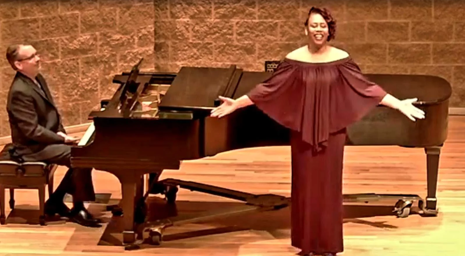 A woman sings on stage in a maroon dress beside a pianist at a grand piano, embodying the spirit of Beacon as Shantée Lewis captivates the audience.