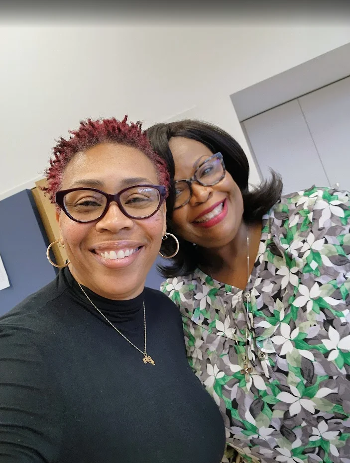 Shantée Lewis and a friend smile and pose together indoors, both wearing glasses and casual clothing—a warm Beacon of friendship.
