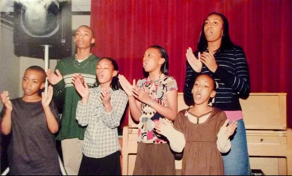 Six children stand in front of a red curtain, singing and clapping like a true Beacon choir, with a piano in the background.
