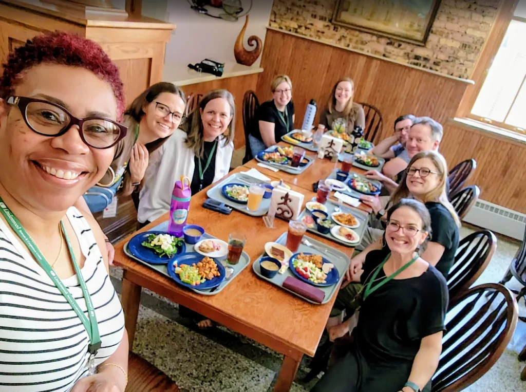 A group of people smiling and eating lunch together at a restaurant, gathered around a wooden table, enjoy lively conversation under the welcoming atmosphere of Beacon, with Shantée Lewis joining in the cheerful gathering.