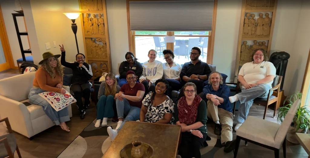 A group of twelve people, including Shantée Lewis, sitting and smiling together in a cozy, well-lit living room—a true beacon of warmth and connection.