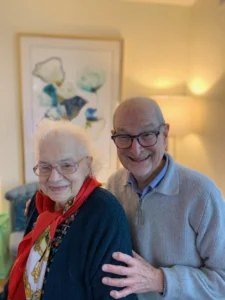 Two smiling elderly people stand together indoors, one with a red scarf, both wearing glasses—capturing a moment of remembering and life in harmony. Photo by Karen Fleischer.