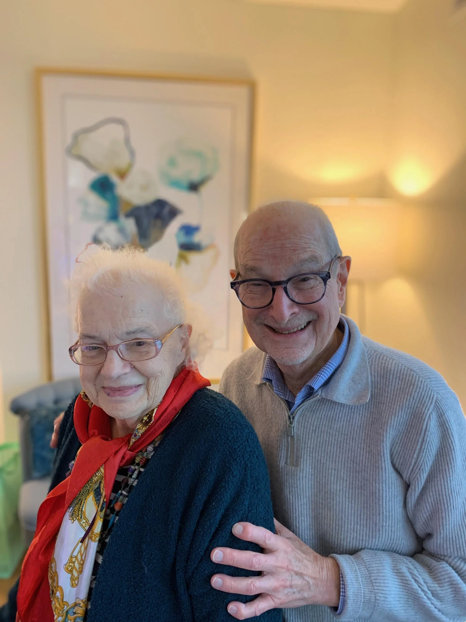 Two smiling elderly people stand together indoors, one with a red scarf, both wearing glasses—capturing a moment of remembering and life in harmony. Photo by Karen Fleischer.