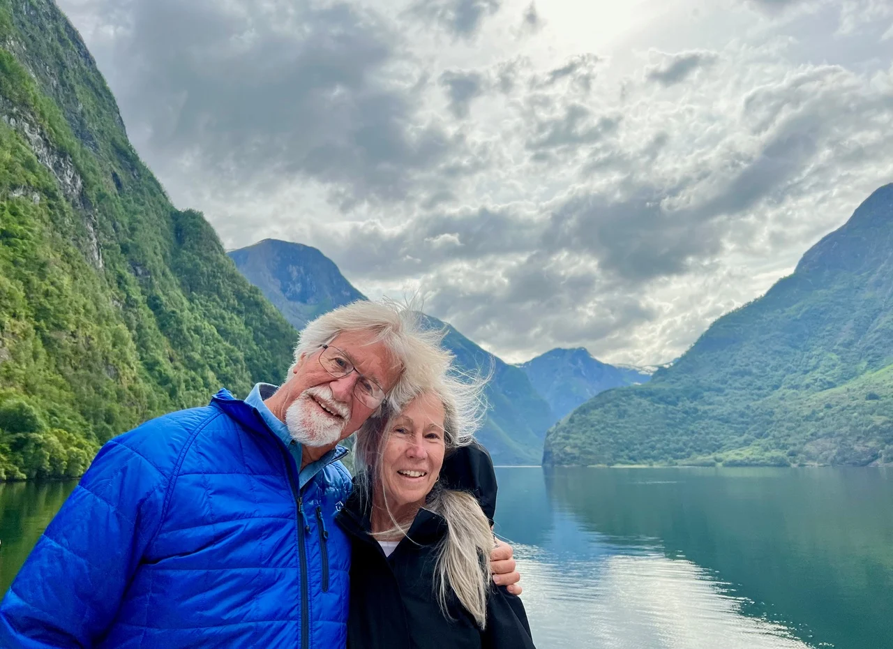 An older couple, proud alumni, smiles by a calm lake, surrounded by green mountains and a cloudy sky—proving with Terry Trautman that there’s never a dull moment in nature’s embrace.