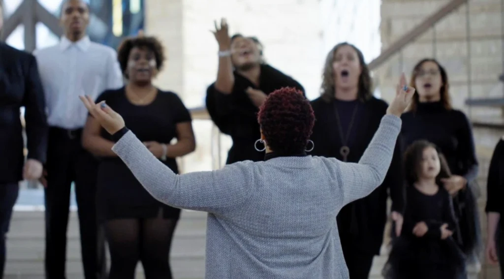 A choir dressed in black sings passionately as a conductor leads them with raised arms inside Beacon, their voices shining like a beacon of unity, reminiscent of Shantée Lewis’s powerful artistic spirit.